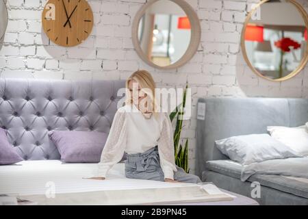 Blonde Frau mit Schmuck am Hals auf dem Bett sitzend. Stockfoto