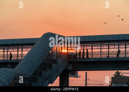 Die Menschen haben sich bei Sonnenuntergang über einer überschlägenden Brücke über eine belebte Straße in der Stadt getuschelt. Niedriger Winkel zur Pansodanbrücke mit unterer Sonne Stockfoto