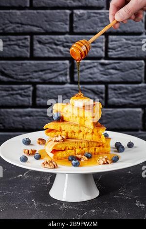 Frau schüttet Honig mit Holzdipper über einen Stapel von Schwammpfannkuchen, die mit Butter, Blaubeeren und Walnüssen bekrönt sind Stockfoto