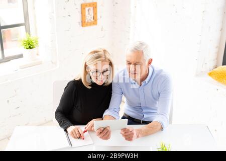 Ein hochwinkeliger Schuss von älteren Ehepaaren, die ihre Finanzen zu Hause berechnen, während sie am Schreibtisch sitzen und ein digitales Tablet verwenden. Stockfoto