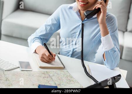 Der Reiseleiter sitzt am Tisch im Büro und beantwortet das Telefon und spricht mit dem Kunden, der Notizen gemacht hat. Er lächelt fröhlich aus der Nähe Stockfoto