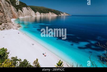 Sommerurlaub. Fteri Strandlagune mit felsiger Küste, Kefalonia, Griechenland. Touristen unter Sonnenschirmen entspannen sich in der Nähe von hellblauem Smaragd Stockfoto