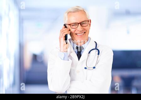 Portraitaufnahme eines älteren männlichen Arztes, der einen Anruf macht, während er in der Arztpraxis steht. Stockfoto