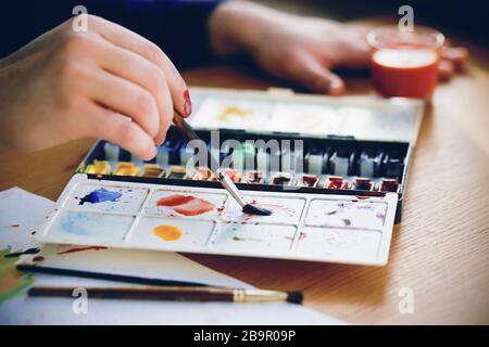 Der Künstler hält einen Pinsel in der Hand, der Farben auf einer Kunststoffpalette mit Aquarellen vermischt, die auf einem Holztisch neben Kunststoffen liegen. Stockfoto