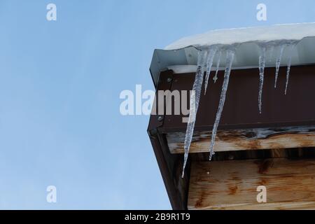 Schmelzende Eiszapfen, die von einer Ecke des Daches hängen, und Wassertropfen fallen auf den Hintergrund des blauen Himmels. Stockfoto