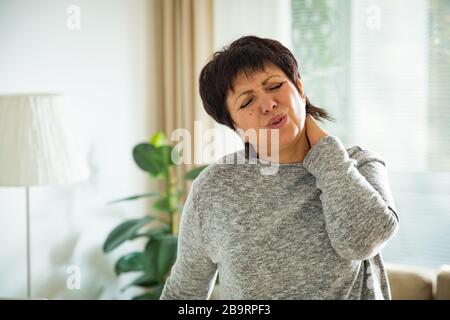 Frau, die zu Hause an Rückenschmerzen leidet. Massieren des Halses mit der Hand, erschöpft fühlen, im Wohnzimmer stehen. Stockfoto