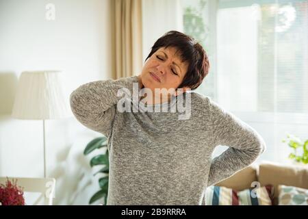 Frau, die zu Hause an Rückenschmerzen leidet. Massieren des Halses mit der Hand, erschöpft fühlen, im Wohnzimmer stehen. Stockfoto