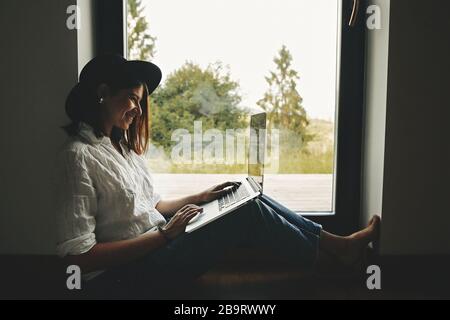 Junge glückliche Frau in Hut mit Laptop, Shopping oder Online-Arbeit von zu Hause aus. Stilvolles Mädchen aus dem Hüftbereich mit Laptop auf dem Boden und großem Fenster mit Blick Stockfoto