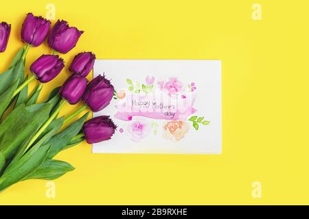 Blumenstrauß aus frischen violetten Tulpen mit Kunsthandwerkspostkarte auf gelbem Hintergrund. Tageskonzept der Mutter. Stockfoto