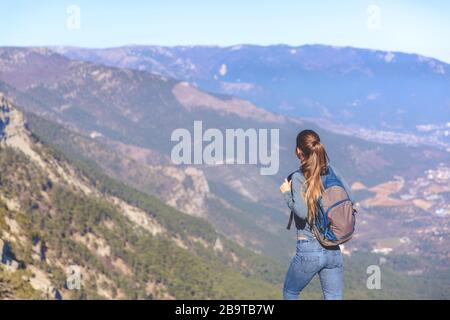 Das junge, schöne Mädchen reist allein im Frühjahr oder Herbst in die Berge, blickt in die Ferne und genießt Natur, Felsen und grüne Wälder, Blick auf die Landschaft, einen Rucksack dahinter und Sportswear, Freiheit und Leichtigkeit Stockfoto