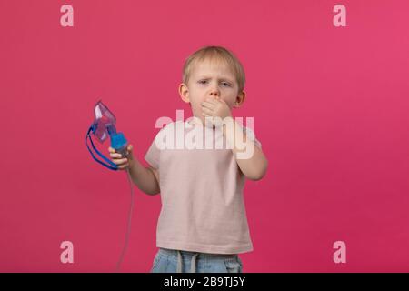 Der blonde Junge hält einen Inhalator in den Händen und hustet, bedeckt seinen Mund mit der Hand. Schuss für Artikel über Erkrankungen der oberen Atemwege, Stockfoto