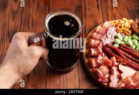 Becher mit Bier und einer Auswahl an Snacks auf einem Holztisch. Wurst, Salami, Schinken, Speck. Bier salzig Snack, Pommes, Wasabi Nüsse, gesalzene Erdnüsse, Croutons. Stockfoto