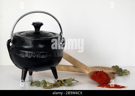 Dreibeiniger schwarzer Topf aus Gusseisen, Holzlöffel und Paprika auf weißem Regal Stockfoto