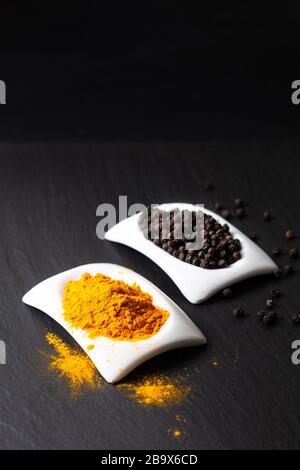 Gesunde Lebensmittel und alternative Medizinzutaten Konzept organisches Turmeriepulver Schwarze Paprika und in quadratischer Keramikschale auf schwarzem Schiefersteinbrett Stockfoto