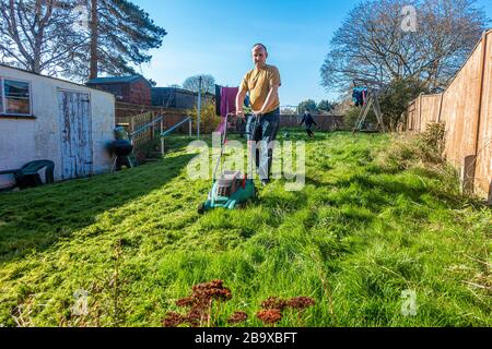 Ein Mann schneidet in diesem Jahr erstmals den Rasen in seinem Hintergarten. Das Gras ist lang und unordentlich. Der Mann schiebt einen elektrischen Rasenmäher Stockfoto