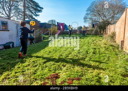 Ein kleiner Junge spielt mit einem Fußball im Hintergarten, während sein Vater einen Rasenmäher in die Ferne drängt. Stockfoto