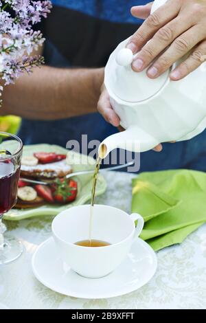 Ein Mann gießt Tee in eine Tasse. Sommerfrühstück im Garten Stockfoto