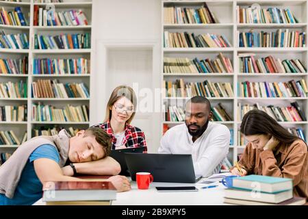 Vorbereitung der Prüfungen. Gemeinsames Projekt. Studenten mit mehreren Rassen, die sich mit Laptop und Buch auf Prüfungen in der Bibliothek vorbereiten. Gutaussehender müde Kerl schläft Stockfoto