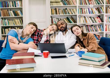 Müde vier gemischte Rennstudenten haben viel zu lernen in einer Bibliothek, lehnen sich gegenseitig und schlafen nach harter Arbeit mit Laptop und Büchern am Tisch Stockfoto