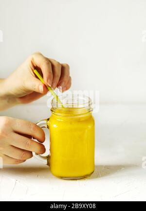 Die Hand der Frau, die ein Glas Banana Mango Smoothie in einem Maurergruß mit einem grünen Strohhalm auf weißem Hintergrund hält Stockfoto