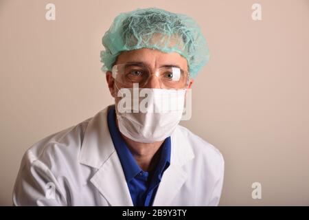 Doktor in seinen Vierzigern mit Gesichtsschutz wie Maske, Brille und Mütze, bekleidet in einer weißen Uniform mit einem Stethoskop, das an seinem Hals hängt und b Stockfoto