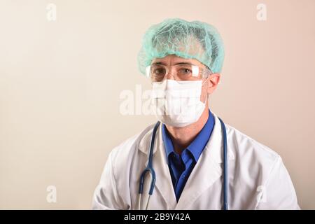 Doktor in seinen Vierzigern mit Gesichtsschutz wie Maske, Brille und Kappe, gekleidet in einer weißen Uniform mit einem Stethoskop, das an seinem Hals hängt und br Stockfoto