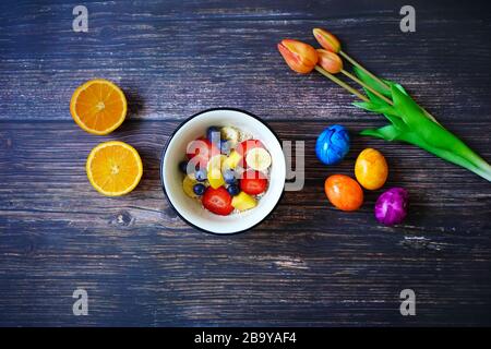 Frühstücksflocken mit frischem Obst (Erdbeere, Banane, Blaubeere, Mango). Halbgeschnittene Orange, bunte Ostereier und orangefarbene Tulpen zur Dekoration Stockfoto