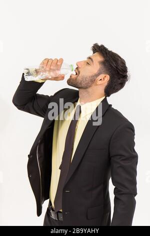 Ein gutaussehender junger Geschäftsmann, der allein steht und Wasser aus einer durchsichtigen Plastikflasche auf weißem Hintergrund trinkt. Stockfoto