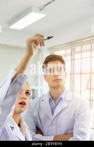 Nahaufnahme: Eine Gruppe junger Wissenschaftler und junger Männer zeichnet Formeln auf einer Glasplatte auf und hat ein orangefarbenes Licht. Stockfoto