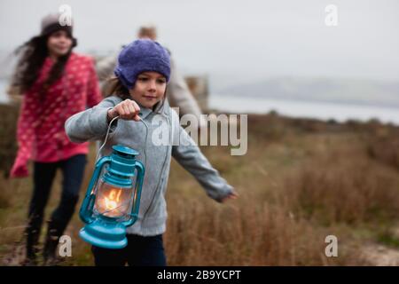 Das junge Mädchen, das eine beleuchtete Laterne trägt, läuft mit ihrer älteren Schwester und einer Frau, die eng folgt, an einem grasbedeckten Ufer entlang. Stockfoto