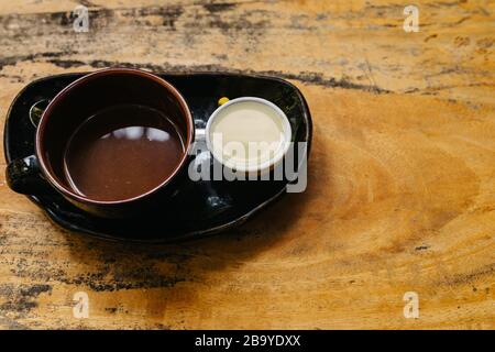 Eine Tasse heißen Espresso in einer dunklen Tasse auf einer schwarzen Untertasse mit Milch separat. Aromatischer Kaffee steht auf einem alten braunen Holztisch. Ort kopieren. Ein guter ST Stockfoto