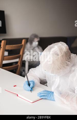 Mädchen im schützenden weißen, lichtdurchlässigen Anzug, blaue Gummihandschuhe, medizinische Maske sitzt zu Hause am Tisch und zeichnet im Skizzenbuch mit Bleistift. Remote-Arbeit von Stockfoto