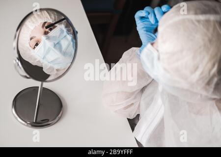 Mädchen im schützenden weißen, lichtdurchlässigen Anzug, blaue Gummihandschuhe, medizinische Maske sitzt zu Hause und malt Wimpern mit Mascara am Spiegel während Stockfoto