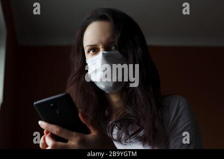 Kaukasisches Mädchen mit dunklem Hörgeräusch in der Arzneimaske, das ihr Telefon hält und besorgt aussieht. Coronavirus (kovid 19) Quarantäne. Stockfoto