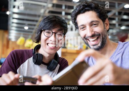 Zwei junge Leute verschiedener Ethnien sitzen an einem Cafeteria-Tisch und machen mit ihren Handys ein selfie, während sie lächeln Stockfoto