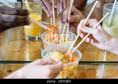 Hände von Menschen verschiedener Ethnien teilen asiatische Speisen in einer Plastikschale mit Stöcken auf einem Holztisch mit Brille von Fruchtsäften Stockfoto