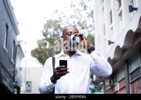 Afroamerikanischer Mann trinkt Kaffee zum Mitnehmen und benutzt sein Telefon Stockfoto