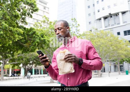 Afroamerikanischer Mann, der sein Telefon benutzt und ein Mitnahme-Sandwich isst Stockfoto