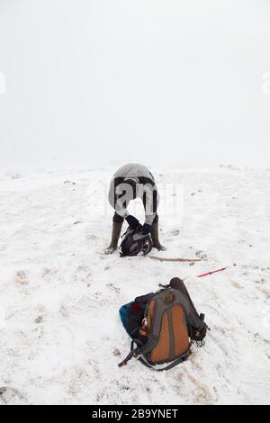 Bergwanderer mit Hut und Gummistiefeln am Oben auf dem Berg suchen in seinem Rucksack und eine andere Oranger Rucksack im Schnee vor ihm Stockfoto