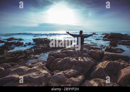 Am Abend seekapern. Felsige Küste bei Sonnenuntergang. Silhouette eines Mannes mit Händen in der Luft am Strand mit Blick auf den magischen Sonnenuntergang Stockfoto