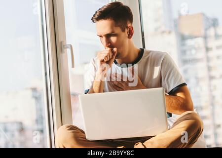 Junger kranker Mann hustet oder niest viel und fühlt sich schlecht an, während er mit dem Laptop auf Runden sitzt. Kranker freier Student hat Atemprobleme während Wor Stockfoto