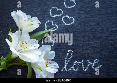 Hintergrund mit weißen Alstroemeria-Blumen oder peruanischer Lilie oder Lilie der Inka mit Herz und Textliebe auf einer schwarzen Kachel Stockfoto