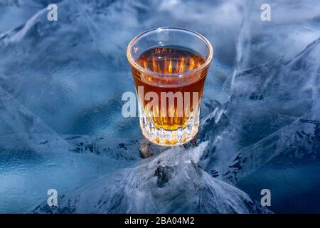 Auf dem Klareis des Sees befindet sich ein Glas Whisky. Gekühlter Whiskey. Blaues Eis mit wunderschönen tiefen weißen Rissen. Draufsicht von der Seite. Stockfoto