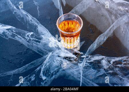 Auf dem gerissenen Eis des Sees befindet sich ein Glas Whisky. Gekühlter Whiskey. Blaues Eis mit wunderschönen tiefen weißen Rissen. Draufsicht von der Seite.