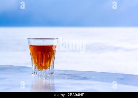 Gekühlter Whisky in einem Glas steht vor dem Hintergrund verschwommener Schnee und Himmel auf Eis. Oranges alkoholisches Getränk in einem Glas. Kopierbereich. Seitenansicht. Stockfoto