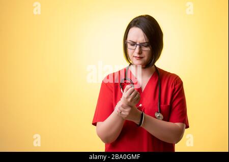 Porträt der schönen Frau Doktor mit Stethoskop mit roten Schruben, hat Handgelenkschmerzen, die auf einem gelb isolierten Hintergrund posieren. Stockfoto