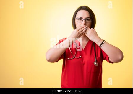 Porträt einer schönen Frau Doktor mit Stethoskop mit roten Schruppen, bedeckt Mund wie Mute-Konzept, posiert auf einem gelb isolierten Hintergrund. Stockfoto