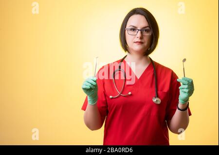 Porträt der schönen Frau Doktor mit Stethoskop mit roten Schruppen, hält in seinen Händen medizinische Instrumente, die auf einem gelb isolierten Hintergrund posieren. Stockfoto