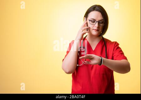 Porträt der schönen Frau Doktor mit Stethoskop mit roten Schruben, Gespräch auf Handy, Smartphone mit Blick auf ihre Uhr posieren auf einem gelben isoliert Stockfoto