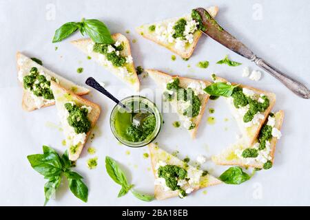 Knusprige Toast (Krostini, Bruschetta) mit Hüttenkäse (Ricotta) und frisch zubereitetem Pesto. Hausgemachte Basilika-Pechto-Soße in einem Glas auf einem hellen Backgroschen Stockfoto